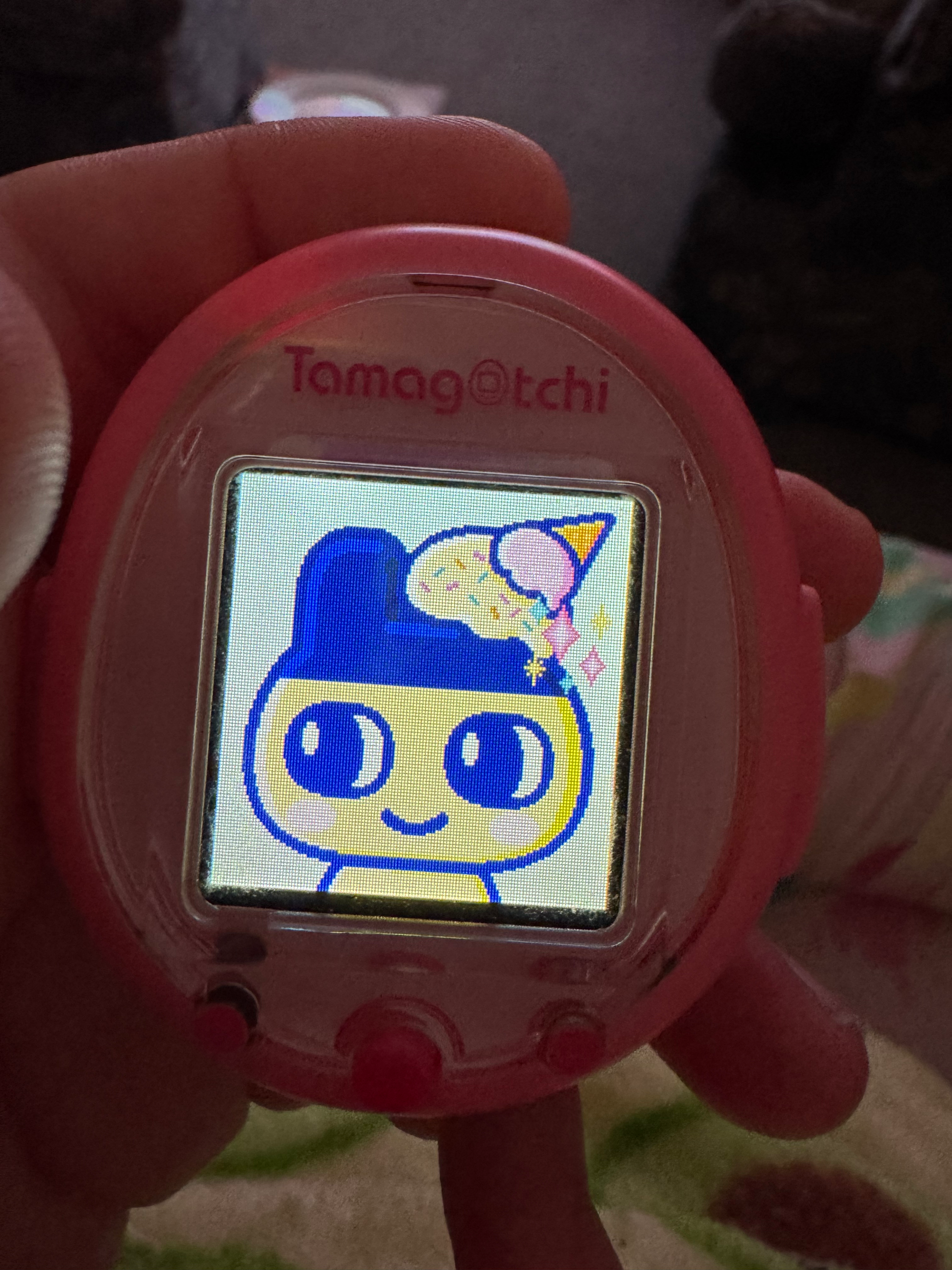 A pink tamagotchi smart with a Mametchi (a yellow dude with blue rabbit ears) and an ice cream on his head. He is smiling, there are sparkles on the photo.