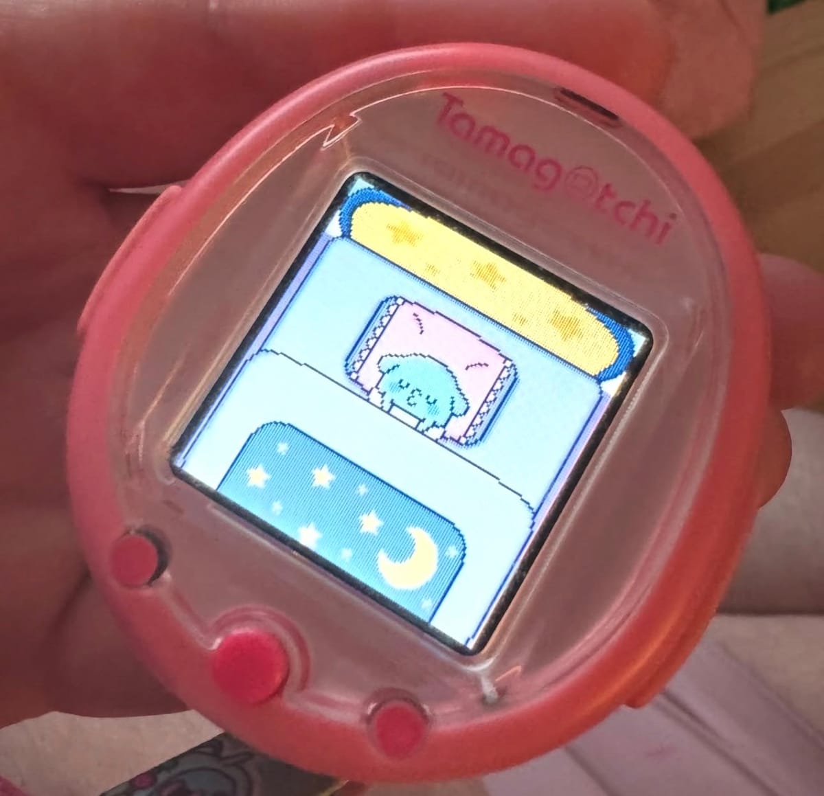 A pink Tamagotchi Smart with a blue character with dog ears in an egg shell tucked into a blue bed with stars and moons on the sheets.